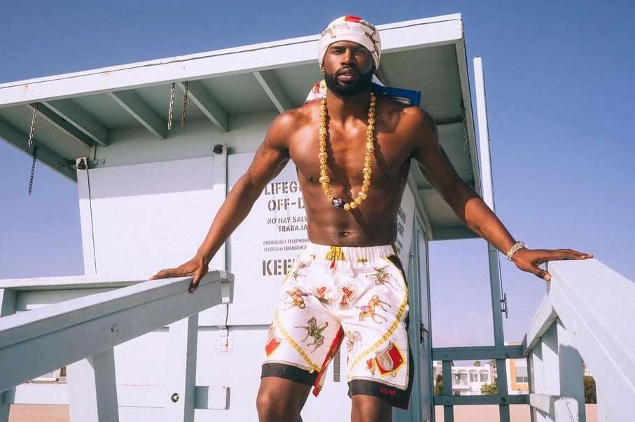 Shirtless African-American man poses near lifeguard tower in patterned shorts, headscarf, and beaded necklace against a blue sky.