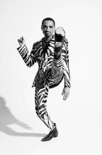 Black and white portrait of a man in zebra-striped suit kicking one leg up in a photo studio, wearing black boots and jewelry.