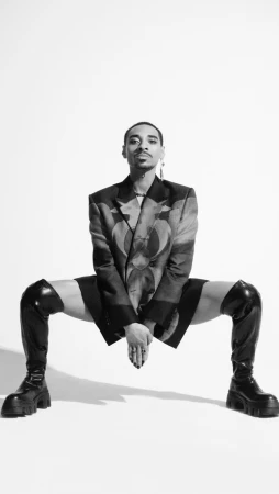 Black and white studio shot of a man in statement blazer, black shorts, and thigh-high platform boots posing against a white backdrop.