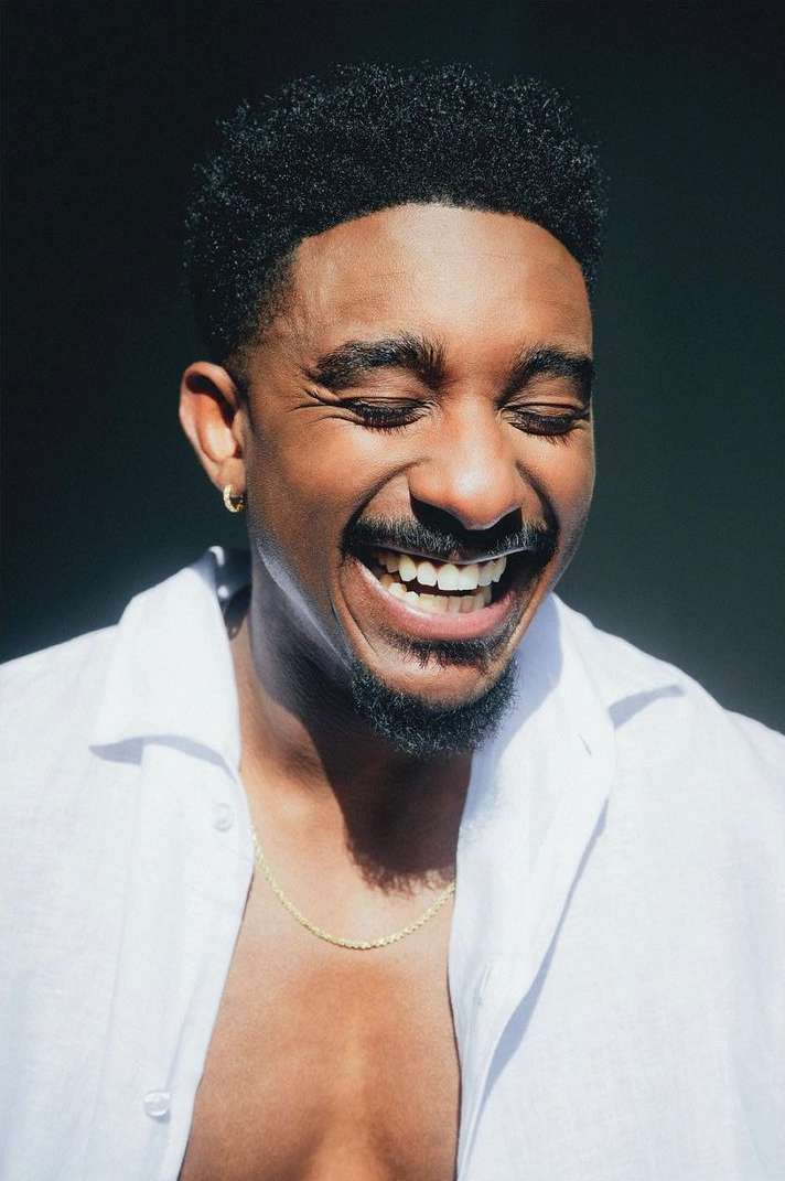 Close-up shot of a smiling Black man in a white collared shirt with a gold chain against a dark background; facial expression of joy.