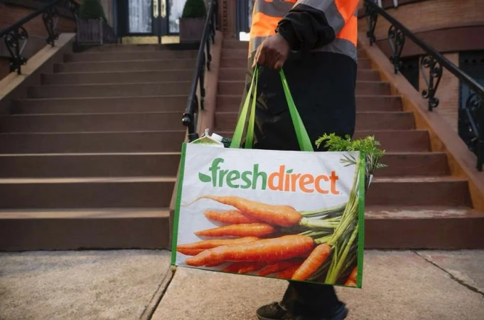 Delivery person carries a Fresh Direct shopping bag filled with carrots up stone steps in front of a brownstone building on a city street.