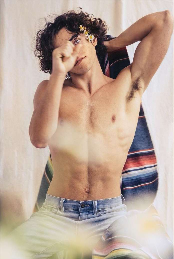 Shirtless man with flowers behind his ear, light jeans. He is seated indoors with a striped blanket in the background.