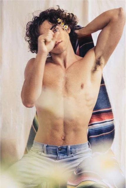 Shirtless man with flowers behind his ear, light jeans. He is seated indoors with a striped blanket in the background.