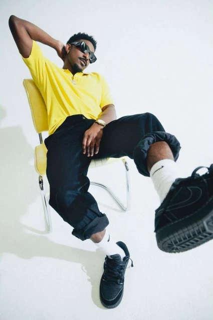 A Black man lounges on a yellow chair in a photo studio, wearing a yellow shirt, black pants, sunglasses, and black sneakers.