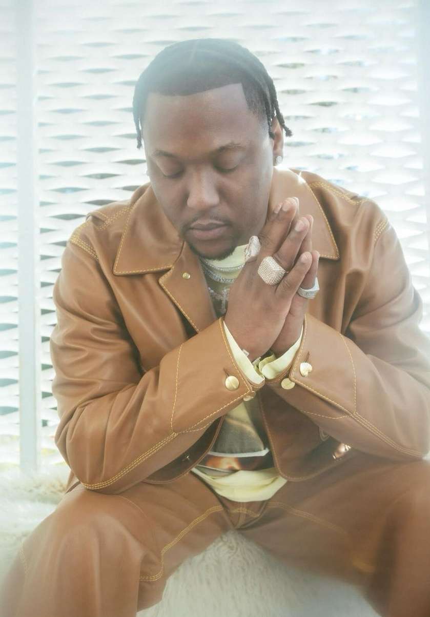 Portrait of a stylish African American man with hands clasped, wearing a tan leather suit in an indoor setting with soft, diffused light.