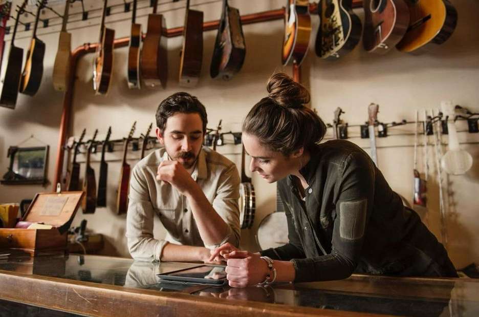 A man and woman stand behind a wooden counter looking at a tablet in a music shop surrounded by guitars and musical instruments.