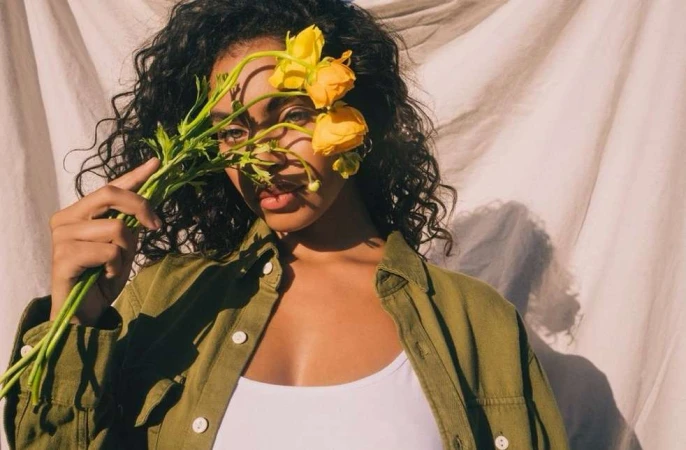 Woman holding yellow flowers in front of her face wearing an olive green shirt against a textured white backdrop.