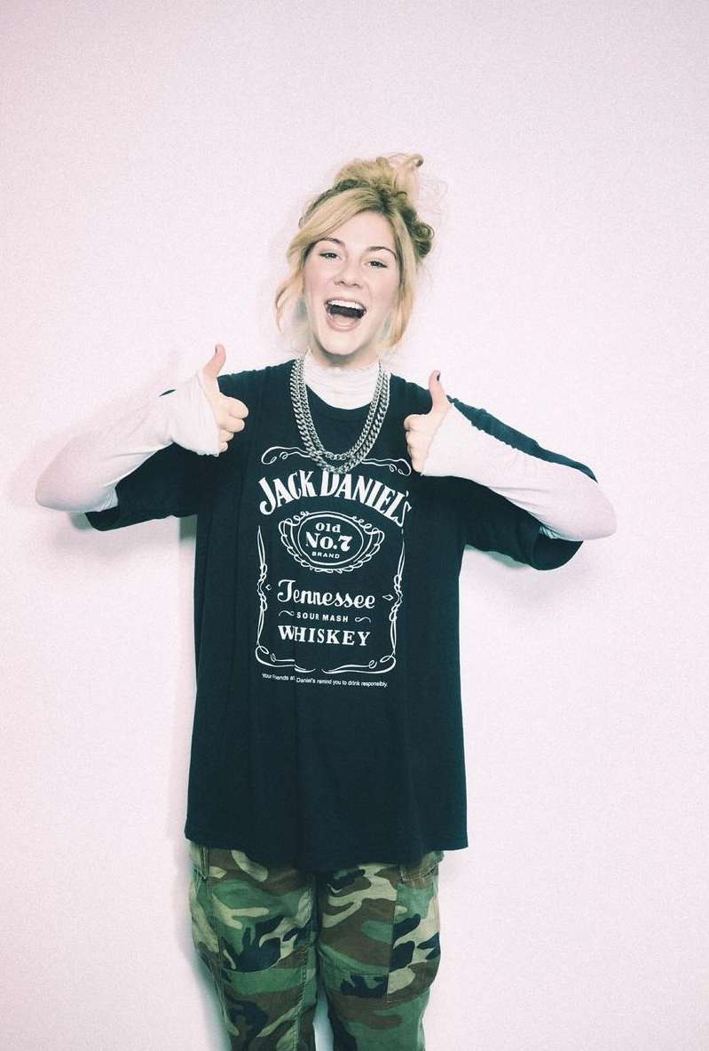 Young woman smiles and gives thumbs up, wearing a Jack Daniel's t-shirt, camouflage pants, and a chain against a white background.