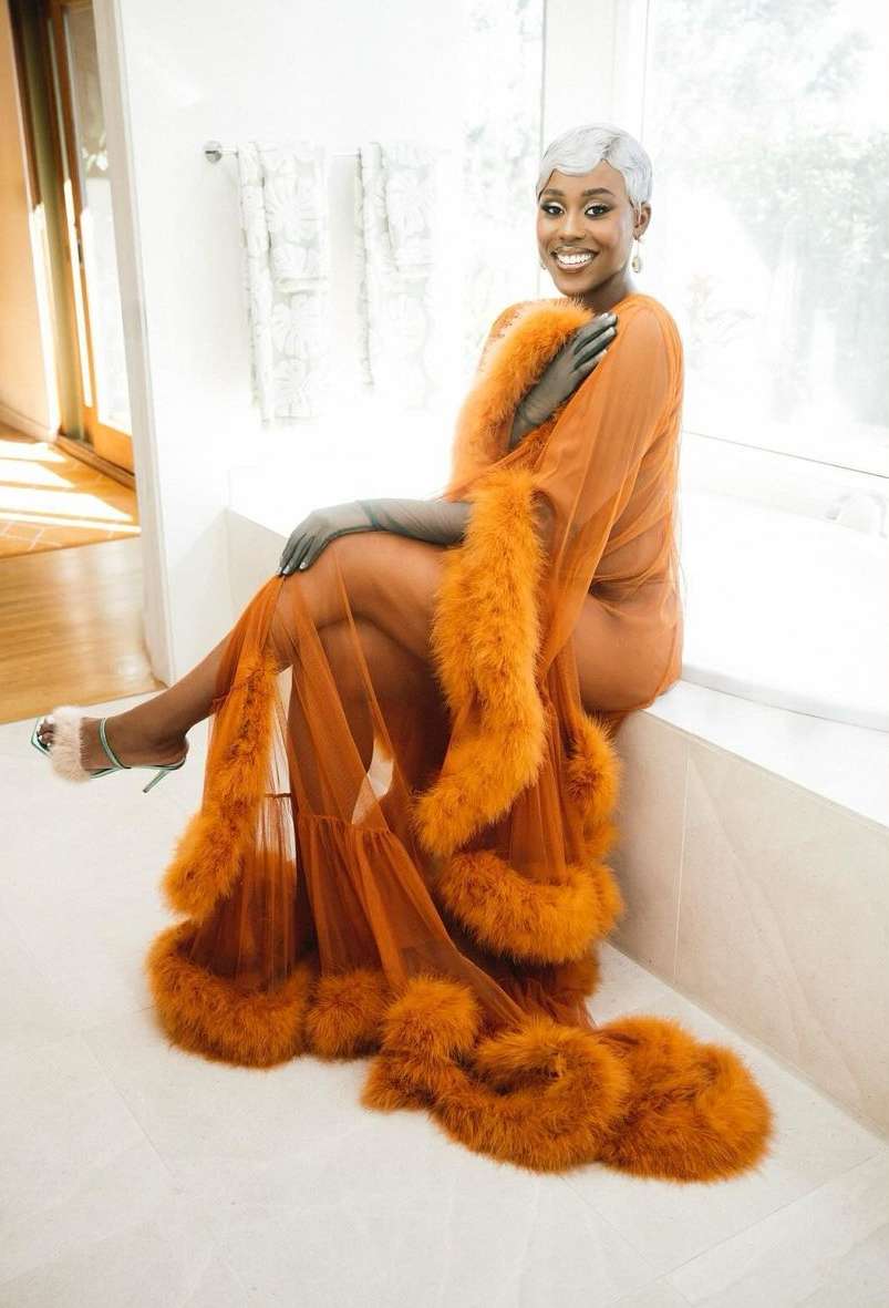 Elegant woman sits in a bathroom wearing a shear orange robe with feather trim. The photo features light and airy ambiance.