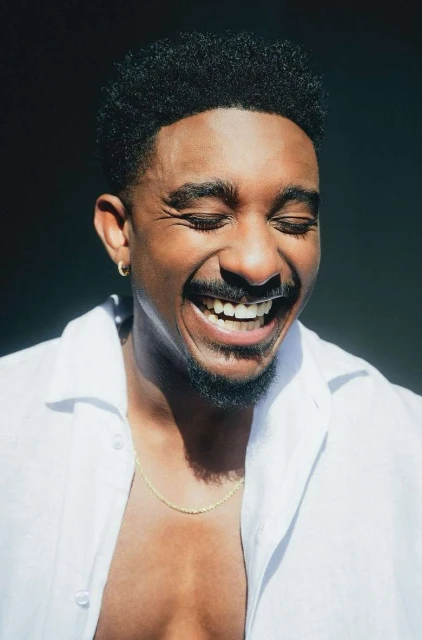 Close-up studio portrait of a smiling adult African American man with a beard wearing an unbuttoned white shirt and gold chain.