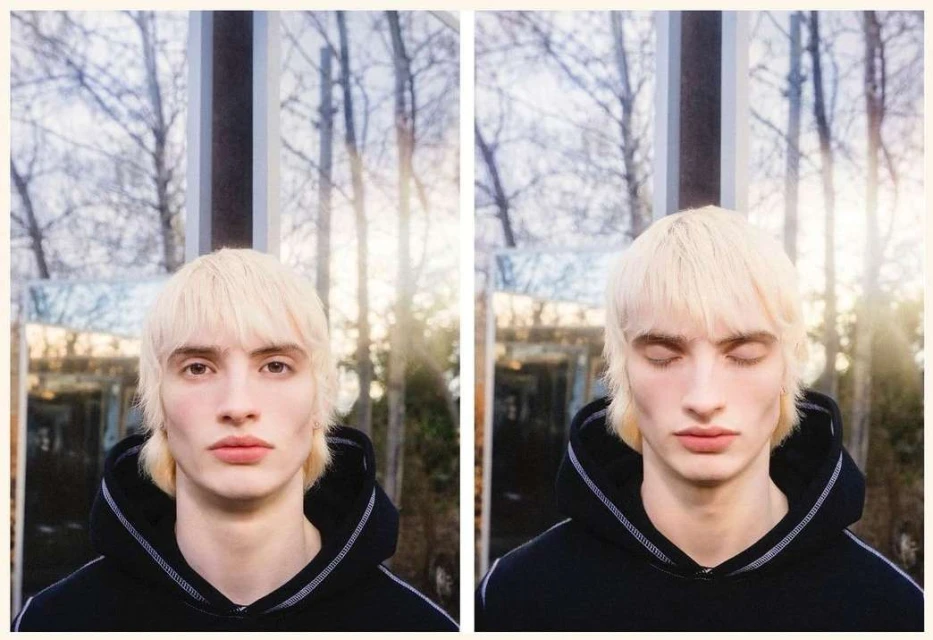 Portrait of a young person with pale blond mullet, wearing a black hoodie, outdoors in a wooded setting with a reflective screen behind.