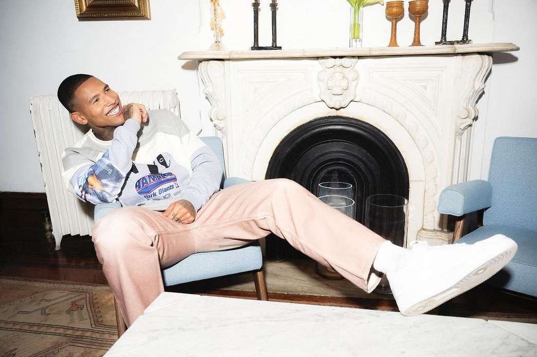 Young man smiling while seated in a light blue armchair wearing a patterned grey sweatshirt and pink trousers