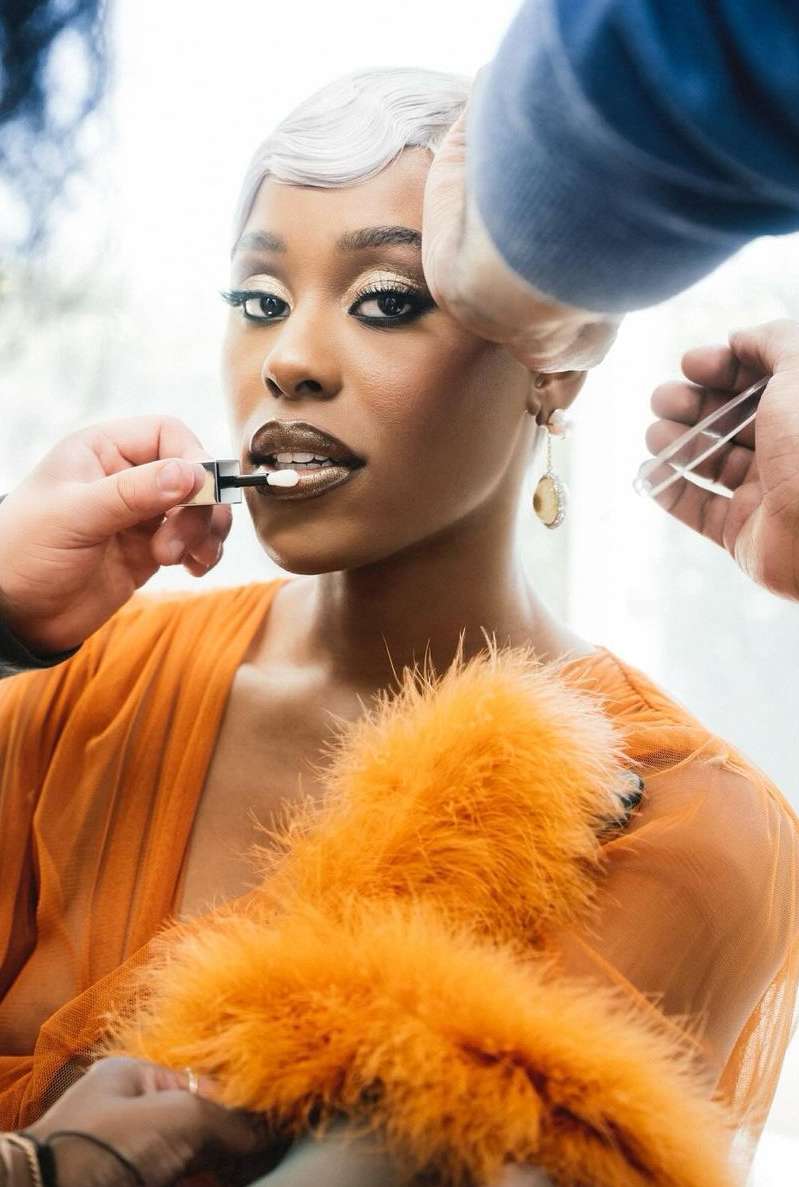 A woman with short silver hair receives a makeup application. She's adorned in an orange top with a feathery boa.