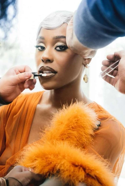 A woman with short silver hair receives a makeup application. She's adorned in an orange top with a feathery boa.