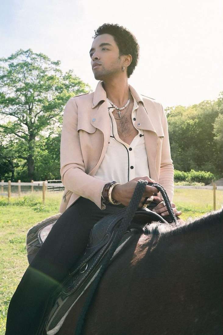 Adult male in jacket riding a dark horse outdoors in a grassy field with trees in the background, looking off into the distance.