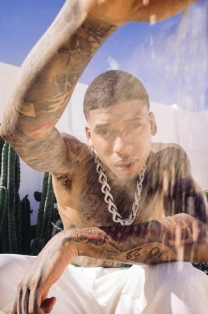 Shirtless man with extensive arm tattoos wearing a chunky silver chain necklace outdoors near cacti.