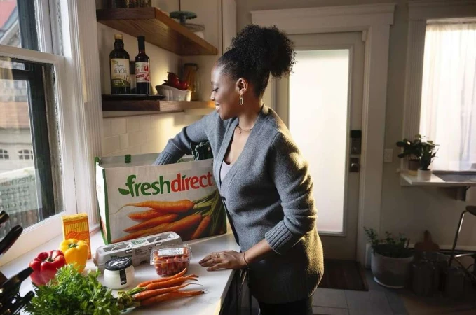 An adult female unloads groceries from a Fresh Direct reusable bag onto a white kitchen countertop indoors with natural lighting.