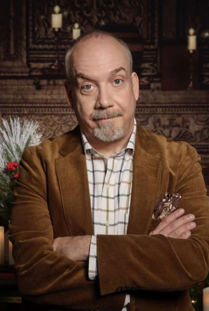 Adult man with beard and mustache wearing brown corduroy jacket, checkered shirt with arms crossed in front of a decorated fireplace.