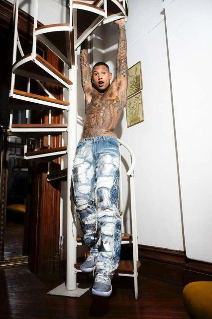Man with tattoos poses on a spiral staircase indoors wearing paint-splattered denim pants and sneakers, framed by dark wooden floors.