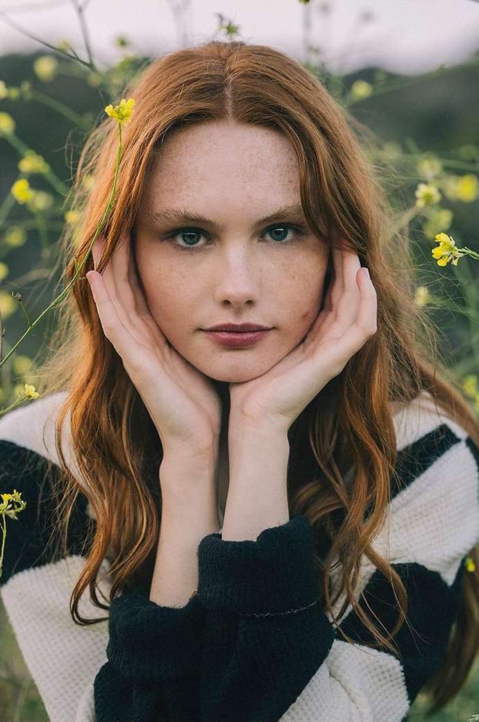 Red-haired woman with freckles poses in a black and white sweater outdoors surrounded by wildflowers during daytime, portrait orientation.