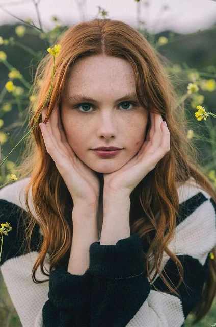 Red-haired woman with freckles poses in a black and white sweater outdoors surrounded by wildflowers during daytime, portrait orientation.
