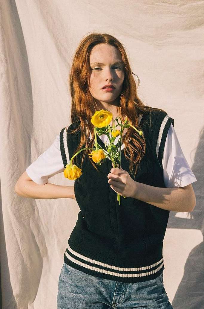 Red-haired woman in a black vest holds yellow flowers against a neutral backdrop, illuminated by warm, diffused sunlight creating soft shadows.