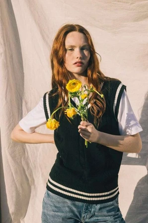 Red-haired female model holding bright yellow flowers wearing a dark sweater vest over white t-shirt and blue denim jeans against fabric backdrop