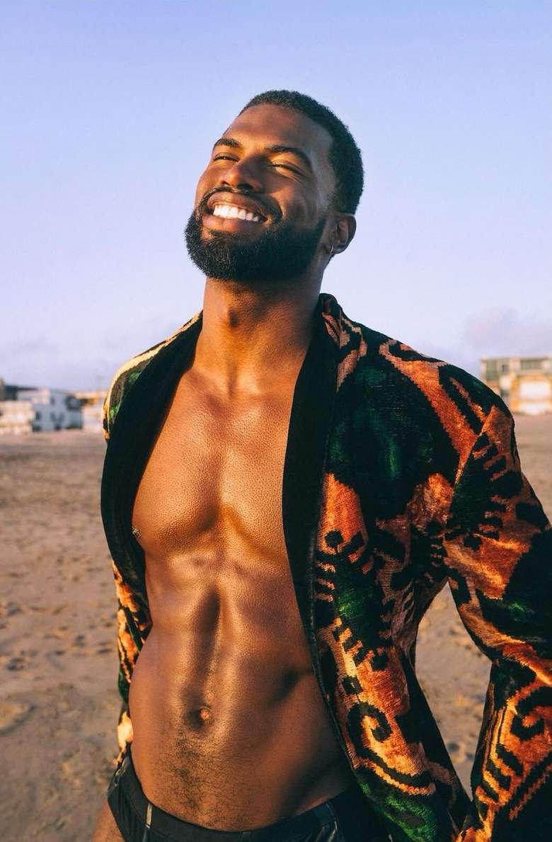 Man on beach smiles wearing patterned velvet jacket. Torso is bare. Sky and sand provide the background. Warm lighting.