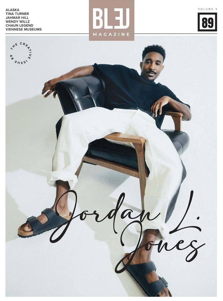 Portrait of a Black man seated in a black leather armchair against a plain white studio background wearing white trousers