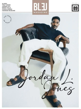 Portrait of a Black man seated in a black leather armchair against a plain white studio background wearing white trousers