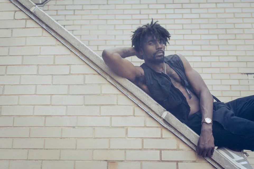 A Black man with dreadlocks wearing a black vest leans against a white brick wall outdoors, posing confidently and looking at the camera.