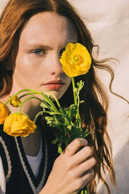 Portrait of a young woman with long red hair, wearing a black sweater vest, holding yellow flowers, against a neutral background.