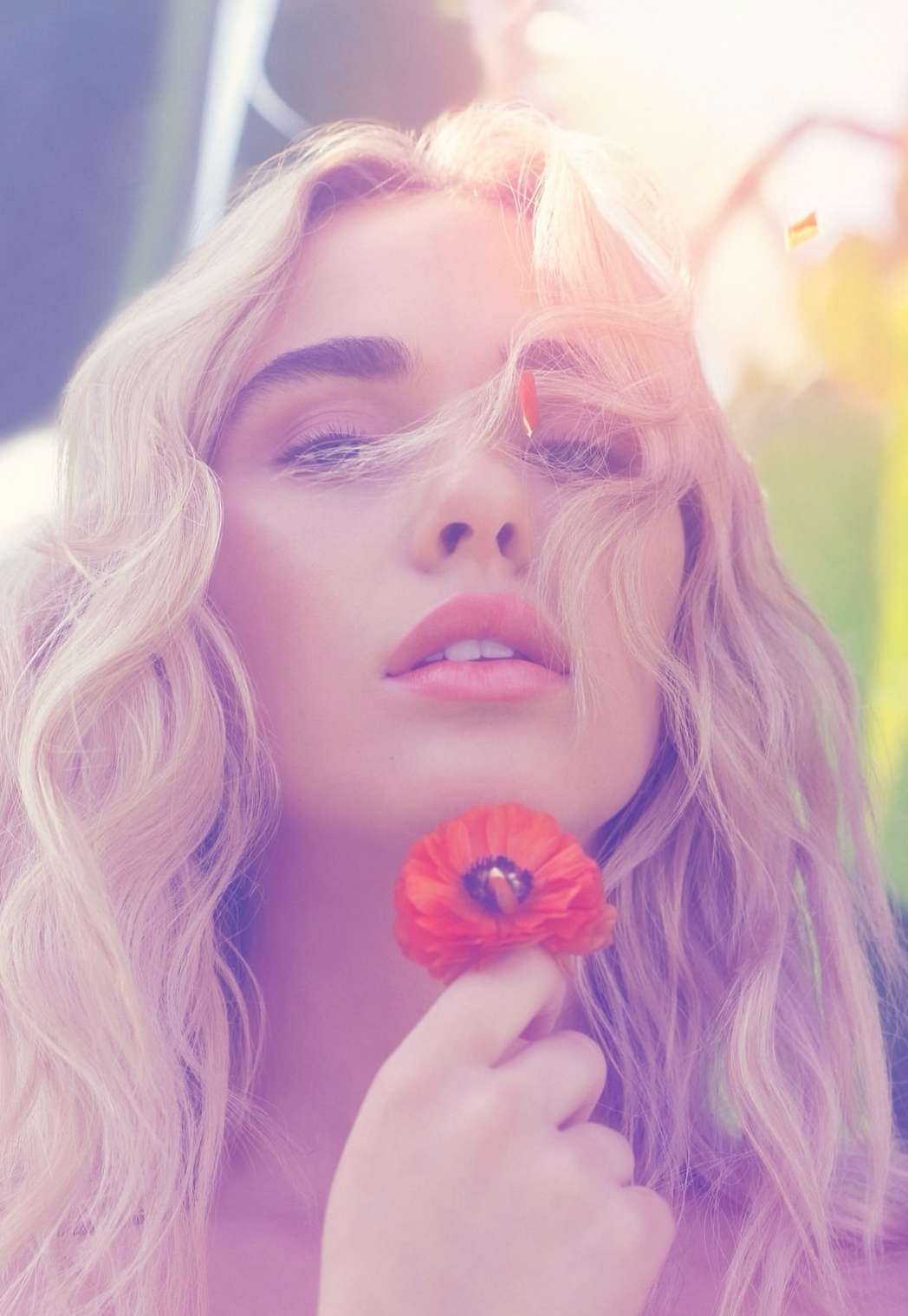 Portrait of a young woman with blonde wavy hair, holding a poppy flower, soft lighting, and a blurred green background, beauty shot.