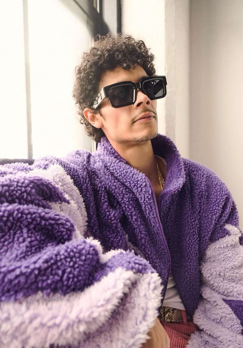 Man wearing a purple fleece jacket and black sunglasses posing near a window in New York