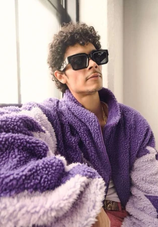 Man wearing a purple fleece jacket and black sunglasses posing near a window in New York