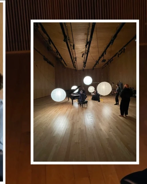 A band rehearses in a large hall with hardwood floors and wood paneled walls, lit by hanging globe lights and stage lights.