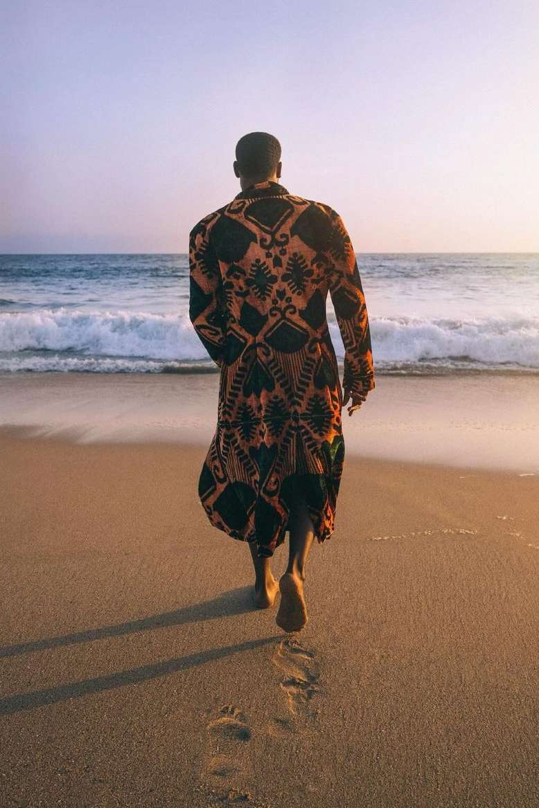 Adult male walks barefoot on sandy beach toward ocean, wearing a patterned robe. Waves roll in under a pastel sky at sunset.