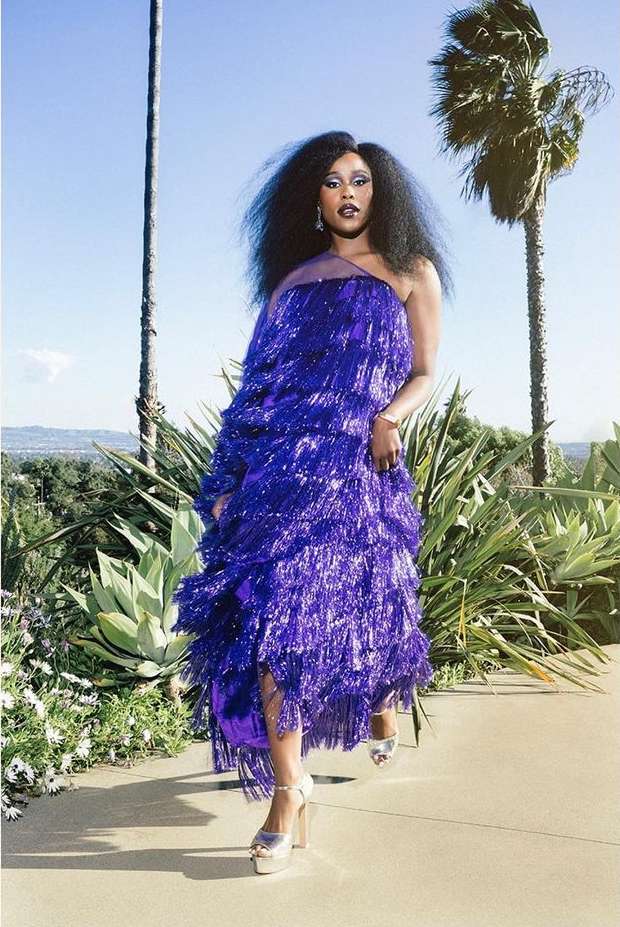 Woman in a shiny blue fringe dress and platform heels poses on a sunny day in a landscape with palm trees and succulents.