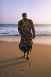 Man walking barefoot away from camera on sandy beach wearing a patterned long coat by photographer Andrew Zaeh
