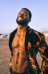 Shirtless black man with beard smiling outdoors on a sandy beach wearing a patterned jacket