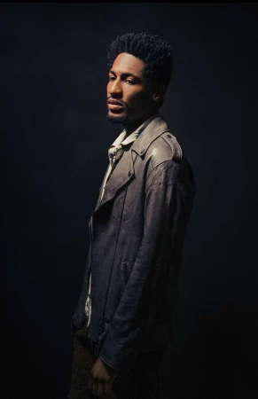 Black man in grey leather jacket posing against a dark textured background photographed by Andrew Zaeh