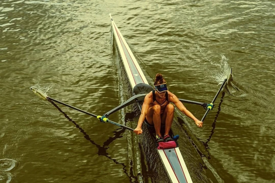 Adult female rowing a scull boat on a river, wearing a visor and athletic clothes; water ripples from oars.