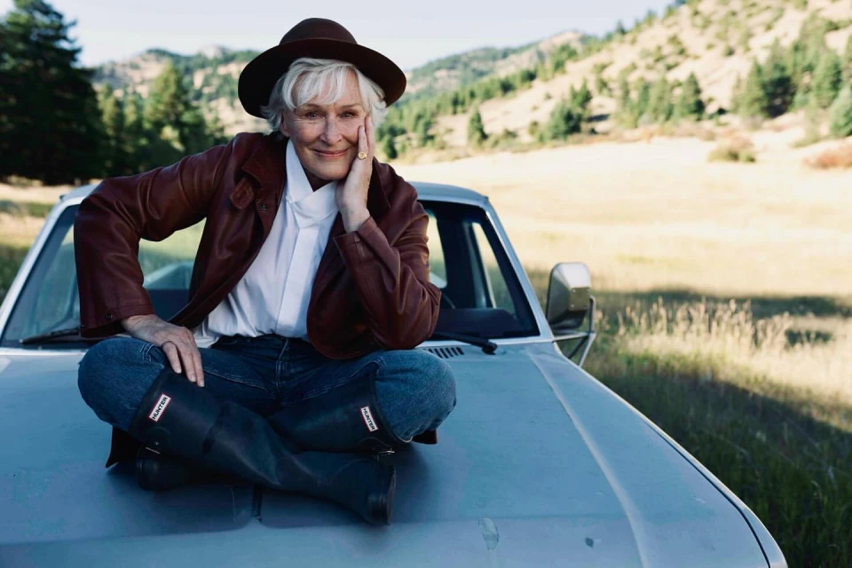 Adult female seated cross-legged on a car hood in front of a mountain landscape, wearing a brown hat, jacket, and black rain boots.