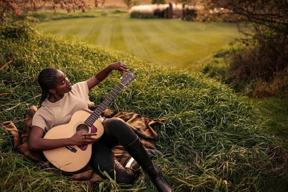 Young Black woman plays acoustic guitar outdoors in a grassy field, wearing a beige shirt, dark pants and boots, relaxed pose.