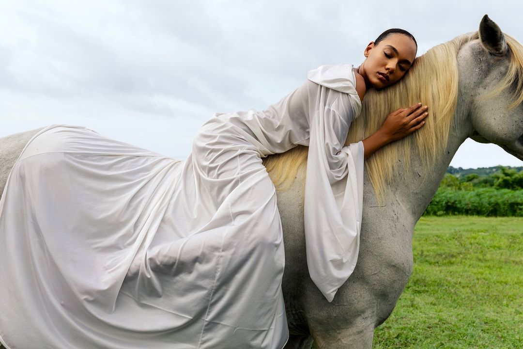 Woman in a white dress laying across a white horse in a grassy field under a cloudy sky, creating a dreamy, ethereal scene.
