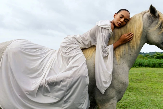Woman in a white dress laying across a white horse in a grassy field under a cloudy sky, creating a dreamy, ethereal scene.