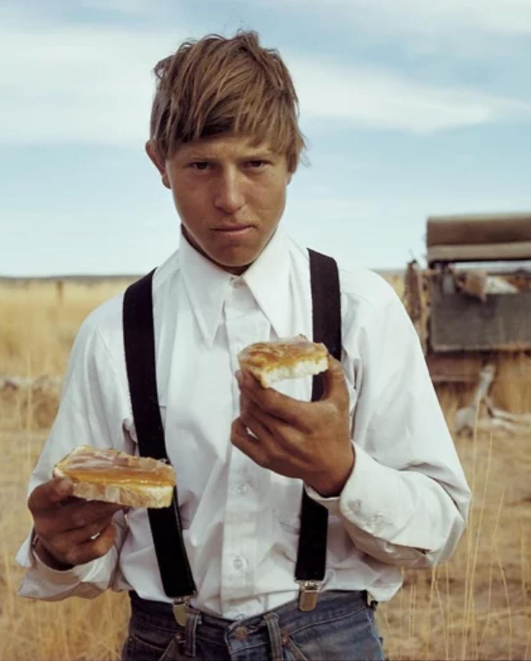 Young adult outdoors holding peanut butter sandwiches in white shirt, suspenders and jeans, against a background of field and sky.