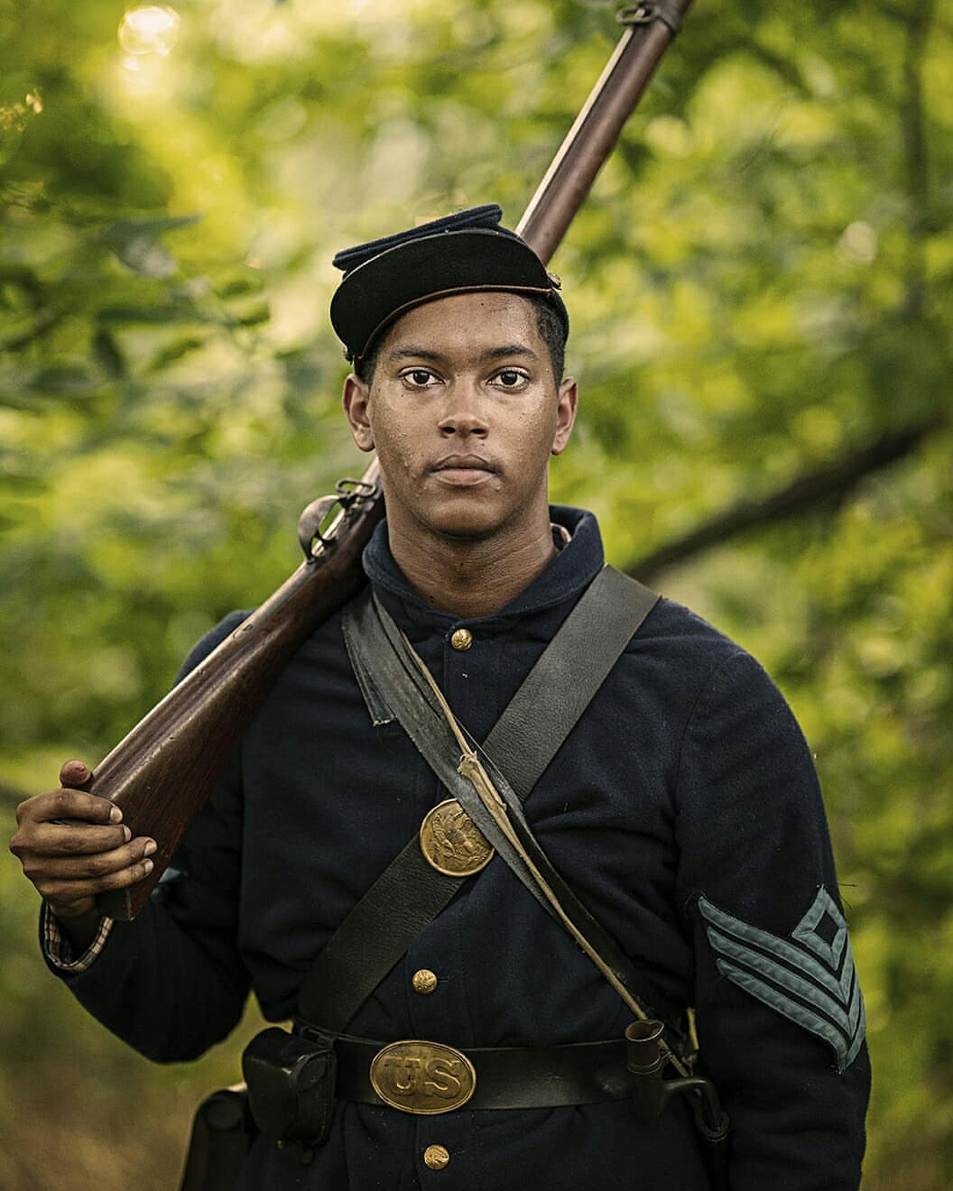Portrait of a young adult in a vintage Union Army uniform with a rifle resting on his shoulder, set against a natural green backdrop.