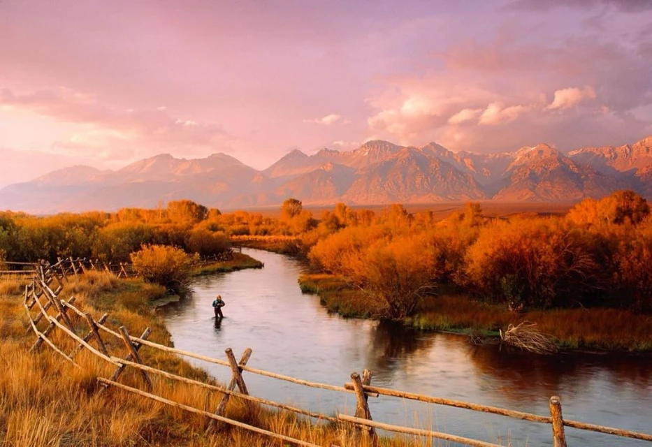 Person fly fishing in a river surrounded by fall foliage and mountains under a pastel sunset sky in an idyllic landscape.