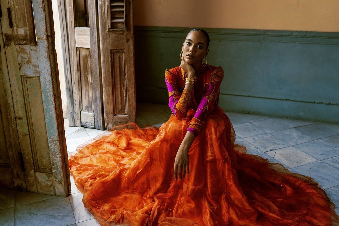 Elegant young woman sits on marble floor in a vibrant orange gown with ornate door in background, captured in a fashion editorial shot.
